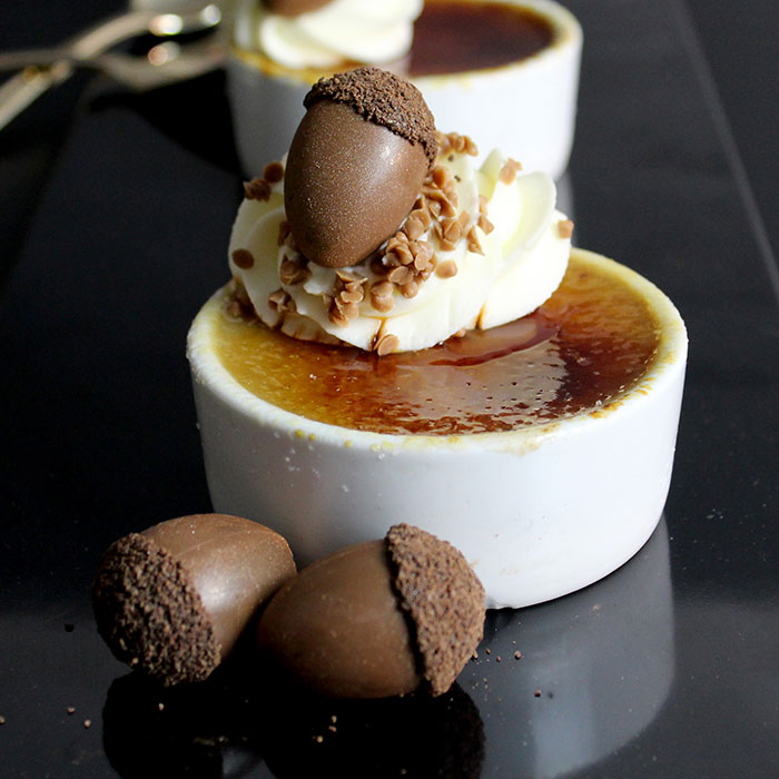 A dish of creme brûlée sits on a black table with whipped cream, butterscotch chips, and a chocolate acorn as decor. Two chocolate acorns sit in front of the dish. 