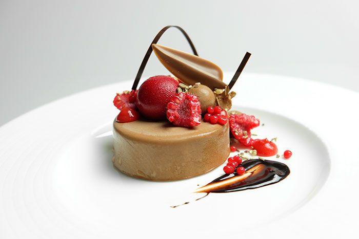 A caramel cake sits on a plate decorated with chocolate ribbons and fresh raspberries. 