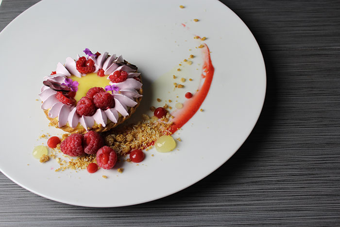 A lemon curd tart sits on a plate with lavender frosting, fresh raspberries, and graham crack crumbs as decor. 