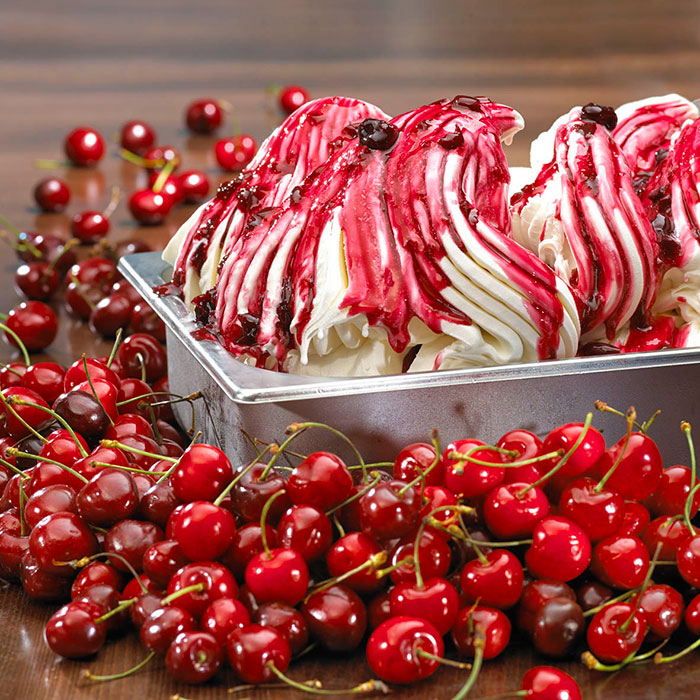 A silver tub of white gelato sits on a dark table topped with a red cherry sauce and surrounded by fresh cherries. 